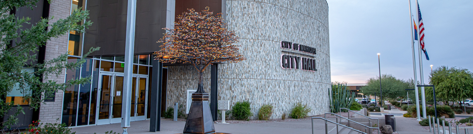 Modern city hall with curved stone facade, large glass entrance, and metal tree sculpture. Flags stand to the right, flanked by lush greenery.