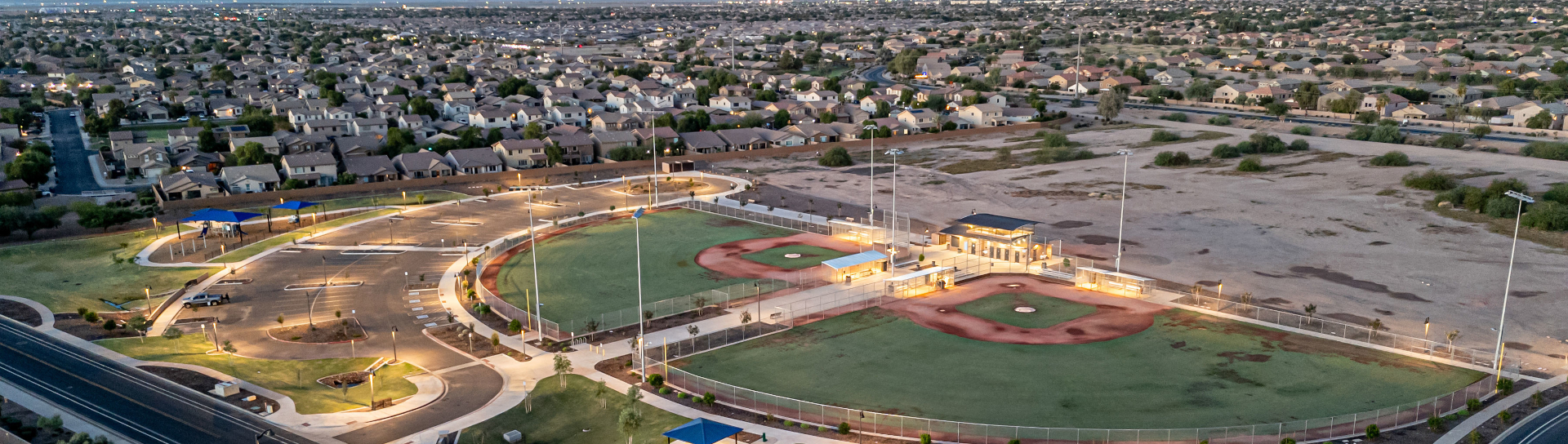 Aerial view of a baseball complex lit by lights at dusk, surrounded by suburban homes and empty lots. The scene conveys a quiet, tranquil atmosphere.