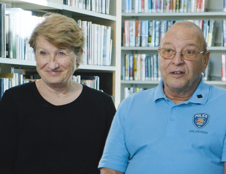 A couple sitting in a library with shelves of books behind them. The woman has short brown hair and a black shirt and the man has short grey hair and a light blue shirt with the City of Maricopa Police Department logo printed on it.