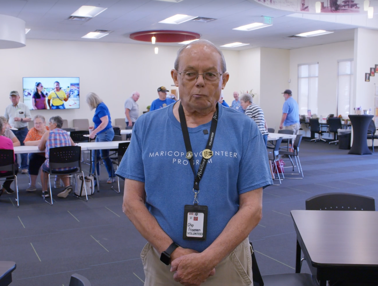 A man standing inside of a community center with people walking around and sitting at tables behind him. He has short grey hair, glasses, and wearing a blue shirt.