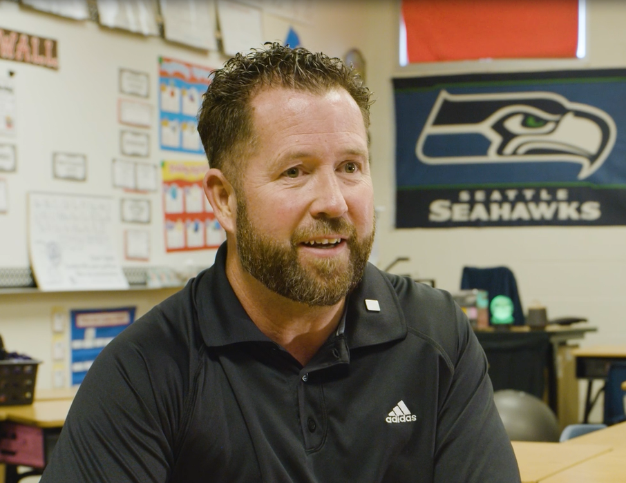 A man with spiked brown hair and a beard sitting in a classroom with a Seattle Seahawks flag in the background.