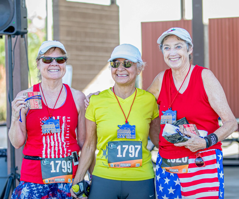 A trio of senior citizens wearing white caps standing and smiling happily while wearing lanyards with racer numbers written on them.