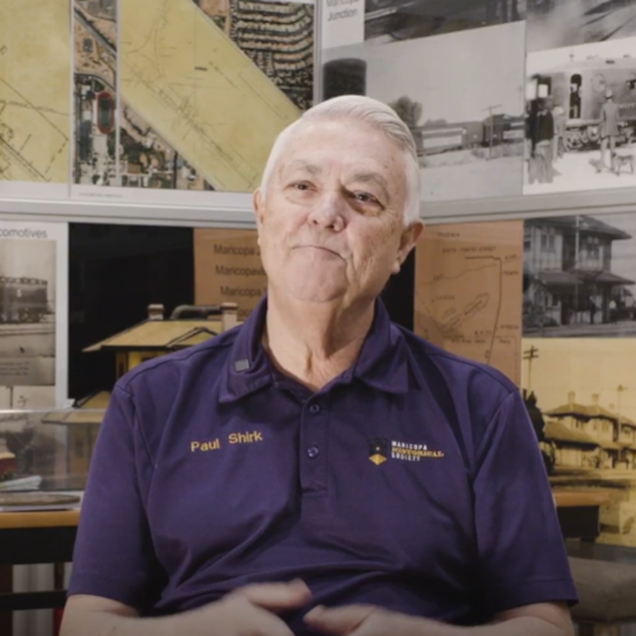 A man sitting in front of a back drop of historical photos smiling with white hair and a purple shirt with the words "Paul Shirk" written on it