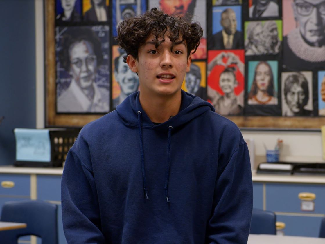 Young kid with brown hair wearing a dark blue hoodie standing in a classroom.