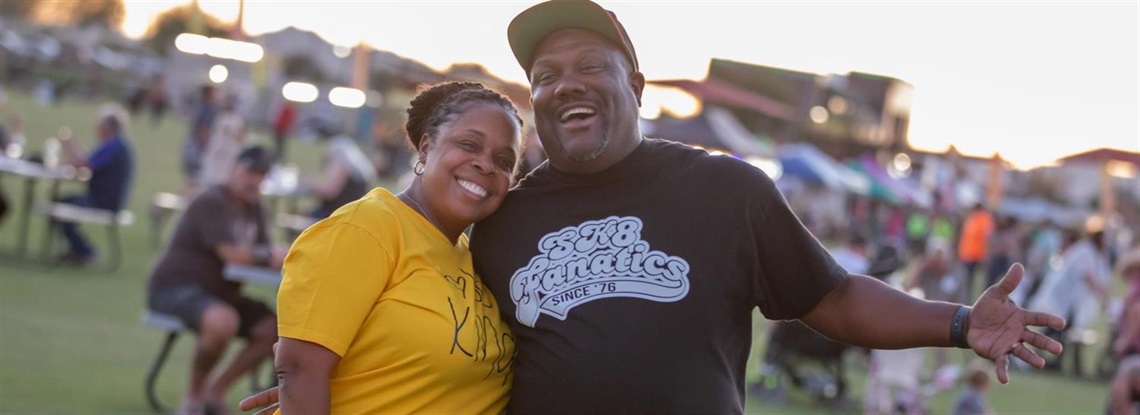 Picture of a woman in a yellow shirt and a man in a black shirt hugging, smiling, and posing in front of the camera in front of a large festival full of people