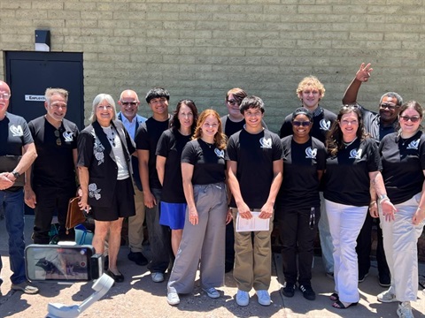 A group of smiling people stand together outside against a brick wall. Most wear matching black t-shirts with a logo. The mood is cheerful and inclusive.