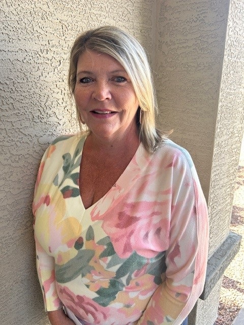 A woman with blonde hair stands against a textured wall. She wears a floral-patterned blouse, smiling softly. Sunlight casts gentle shadows, creating a warm tone.