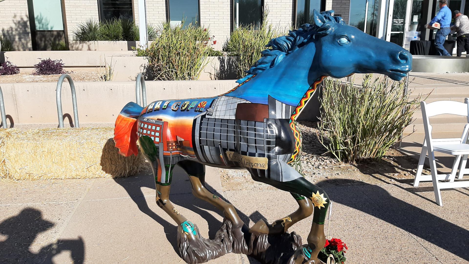 A vibrantly painted horse sculpture stands on a path, featuring urban scenes, geometric patterns, and a bright blue head. Plants and a straw bale are in the background.