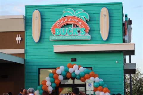 Turquoise building facade of Bahama Buck's, a shaved ice company. Features a logo with surfboards and a festive archway of teal, orange, and white balloons.