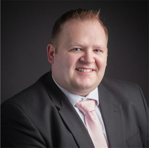 Smiling person in a dark suit and light pink tie, posing against a dark background. The tone is professional and approachable.