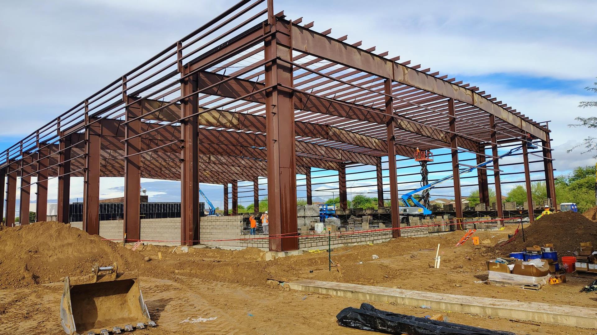 A large construction site with a steel framework for a building. Workers in helmets are seen working. The sky is partly cloudy, and equipment is scattered around.
