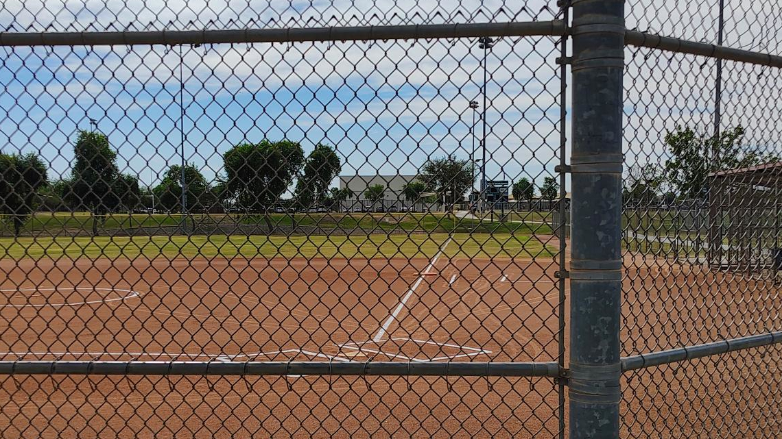 A view through a chain-link fence onto a baseball field, with a clear blue sky and some clouds overhead. The scene is calm and empty, suggesting anticipation.