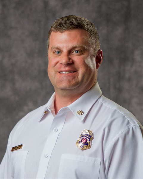 A man in a white uniform shirt with fire department badges poses confidently against a gray backdrop, conveying professionalism and authority.