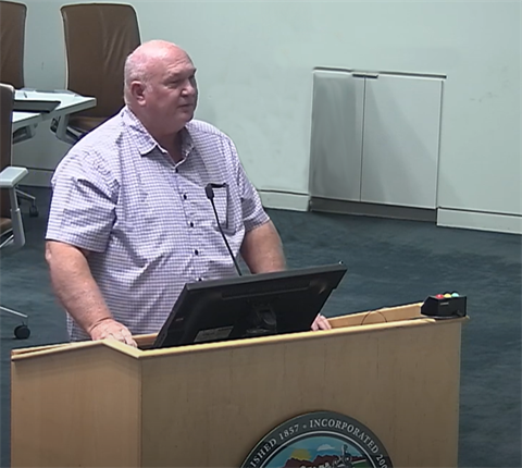 A man in a checkered shirt speaks at a podium with a microphone in a conference room. He stands confidently, with chairs and a cabinet in the background.