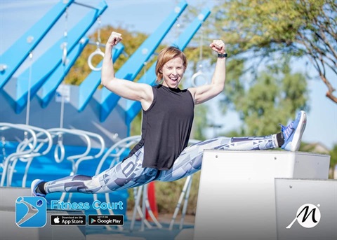 A woman performing a split leap over outdoor fitness equipment, smiling and flexing her arms, exuding strength and energy. Bright, sunny day with trees and blue apparatus in the background. Fitness Court logo and app icons visible.