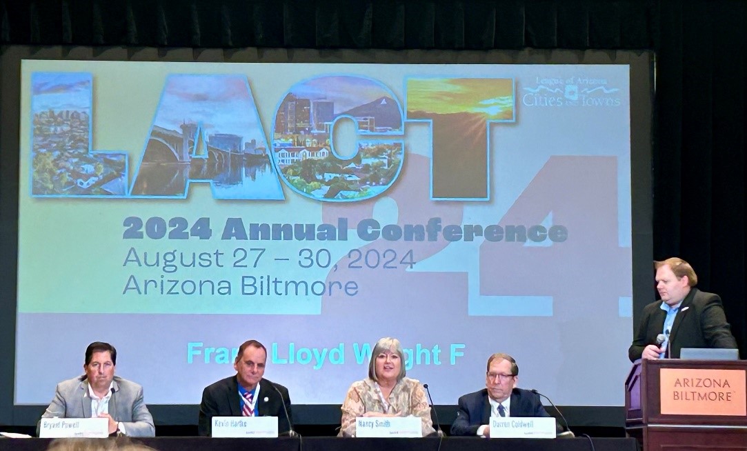 Five people sit at a conference panel table with a projection screen behind them displaying 