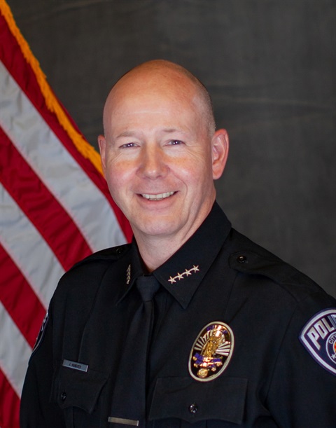 A smiling police officer stands in uniform with badges, in front of the American flag. The atmosphere is formal and respectful.