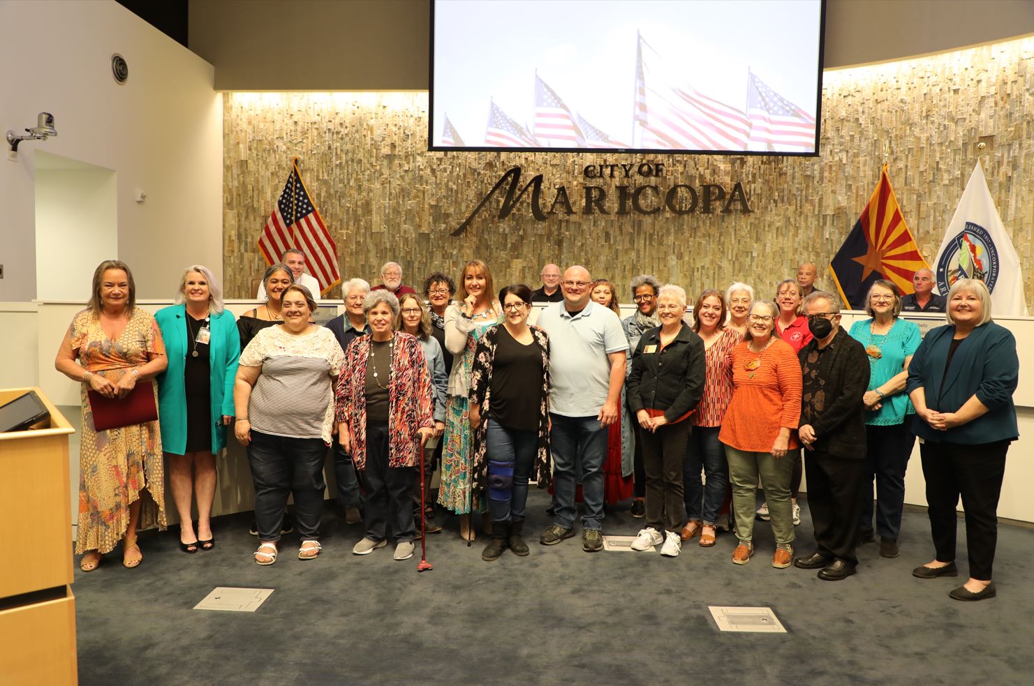 A diverse group of people poses smiling in a city council room. Behind them are U.S., Arizona, and city flags with 