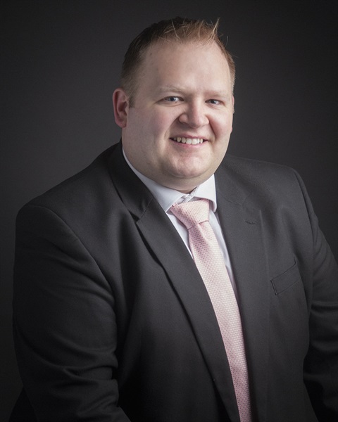 A smiling man in a black suit with a light pink tie poses against a dark background. The tone is professional and welcoming.