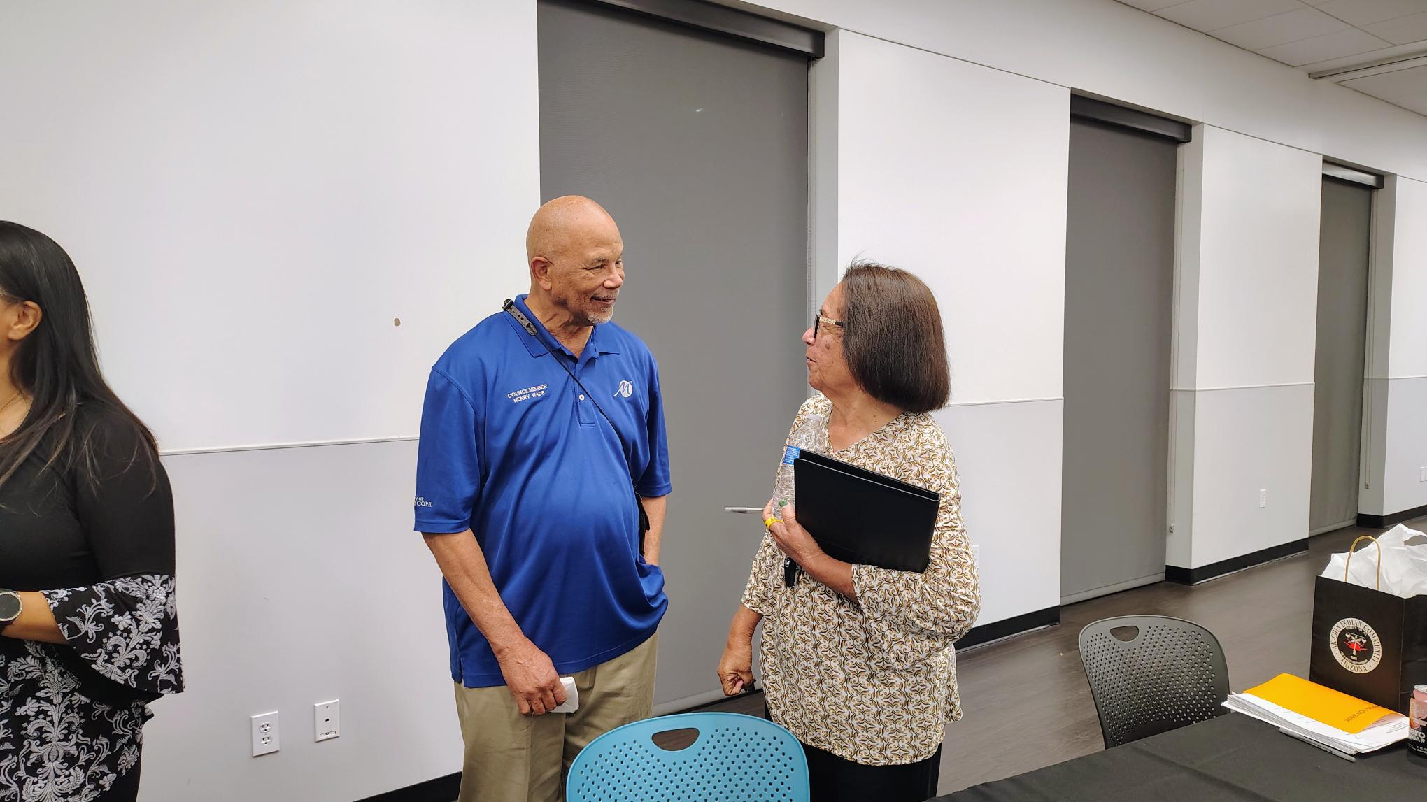An older man in a blue shirt and an older woman with glasses holding a folder are engaged in conversation in a conference room setting.
