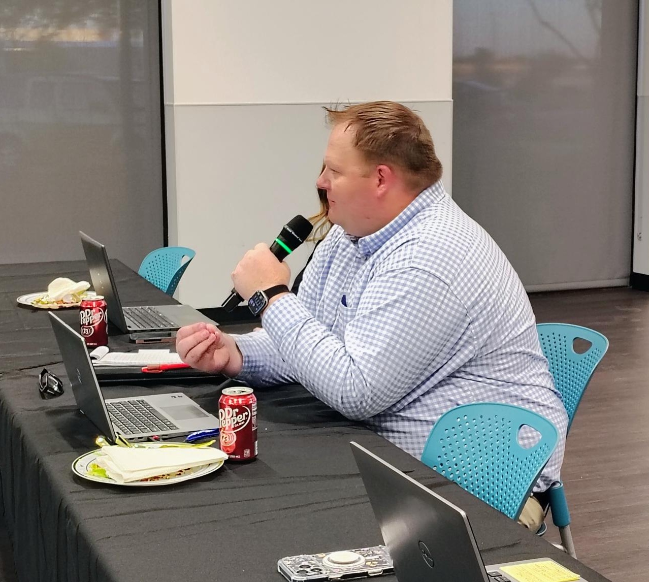 A person in a blue checkered shirt speaks into a microphone while seated at a table with laptops, Dr Pepper cans, and plates.