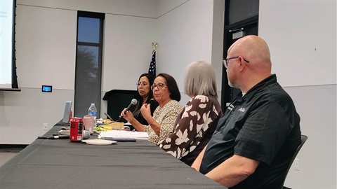 Four people sit at a conference table in a meeting room, with one woman holding a microphone while speaking. An American flag is visible in the background.