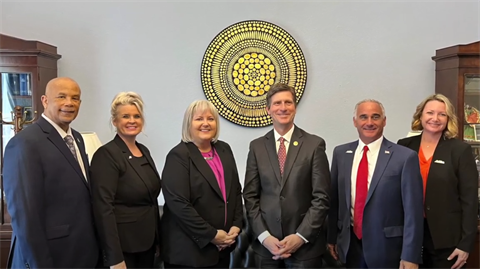 A group of six people in business attire stand smiling in front of a decorative circular wall piece. The setting conveys a formal and positive tone.