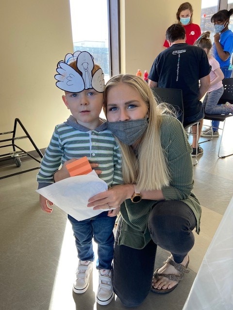 A young child wearing a handmade turkey hat stands next to a smiling woman in a mask. They are indoors, surrounded by people. The mood is cheerful.