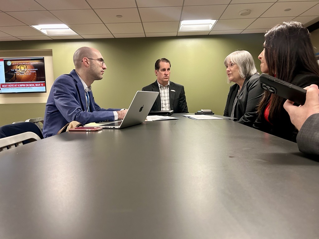 Four people in formal attire sit at a meeting table with laptops and papers, engaged in a discussion. A digital screen displays news in the background.