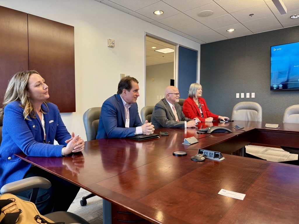 Four people in professional attire sit at a conference table in a meeting room, attentively watching a wall-mounted screen. The atmosphere is focused.