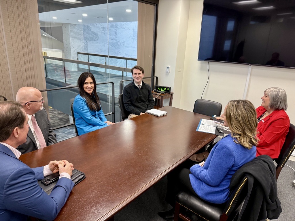 A group of six people in business attire sits around a wooden conference table in a modern, glass-walled meeting room. They are engaged in conversation.