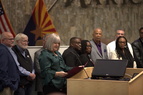 A group of people stand together at a podium indoors, with one woman reading from a document. Flags are displayed in the background, creating a formal setting.