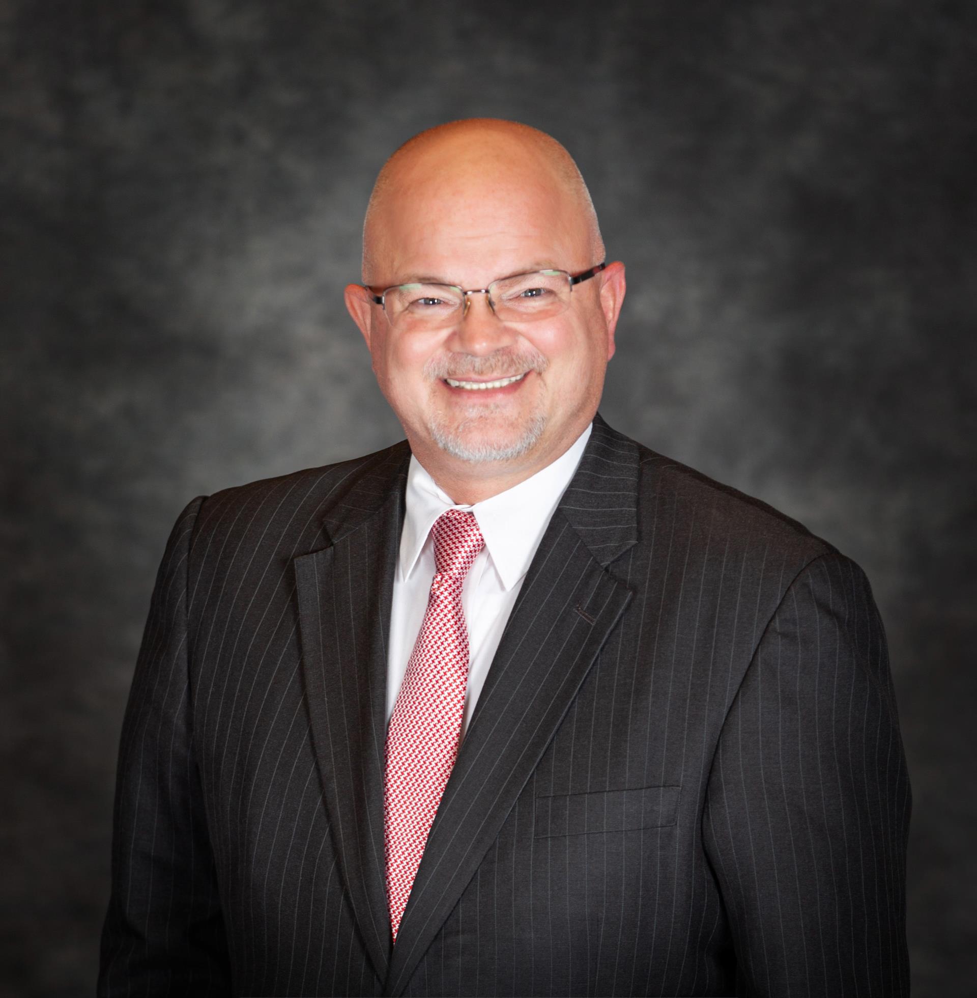 A smiling man in a pinstripe suit and red tie poses against a dark gray background. He exudes confidence and professionalism.