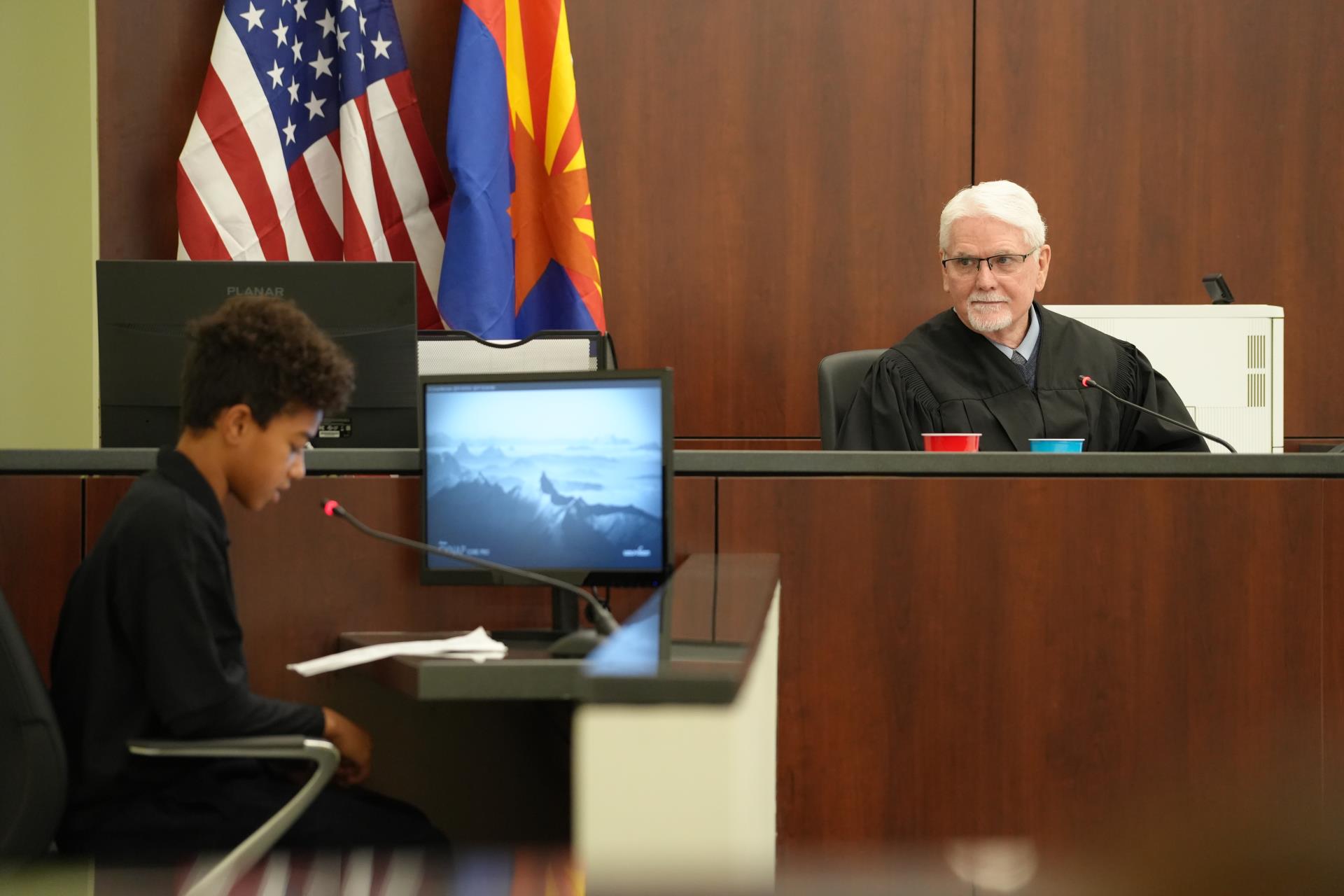 A young person reads in court, facing a judge in robes. U.S. and state flags are behind the judge. The scene is formal and serious.