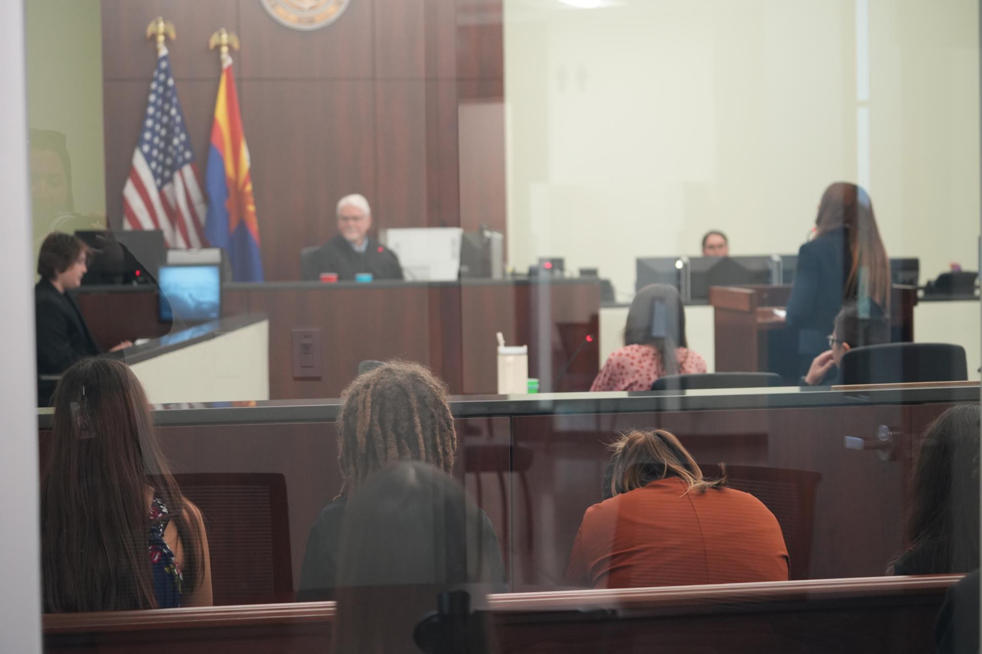 A courtroom scene with a judge, flags behind, and people seated. One person leans forward at a desk. A somber tone is conveyed through the image.