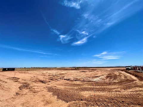 Vast, cleared construction site under a bright blue sky with wispy clouds. Dirt tracks are visible, and a truck is parked in the distance, suggesting development.
