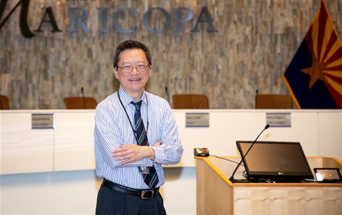 Smiling man in a striped shirt and tie stands confidently with arms crossed in a council chamber. Behind him, a podium and Arizona flag are visible.