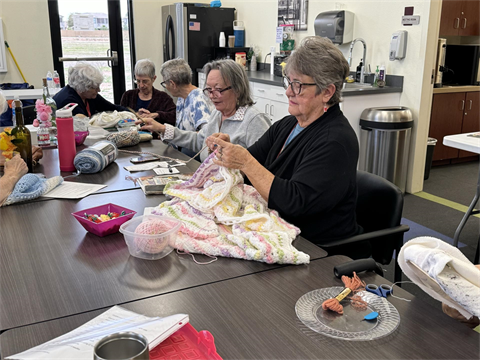 A group of six women sit around a table engaged in knitting. The table is cluttered with yarn, scissors, and patterns. The room has a cozy, focused atmosphere.