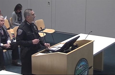 A police officer stands at a podium speaking in a conference room. Behind him are seated attendees, including a man in a suit. The mood is formal.