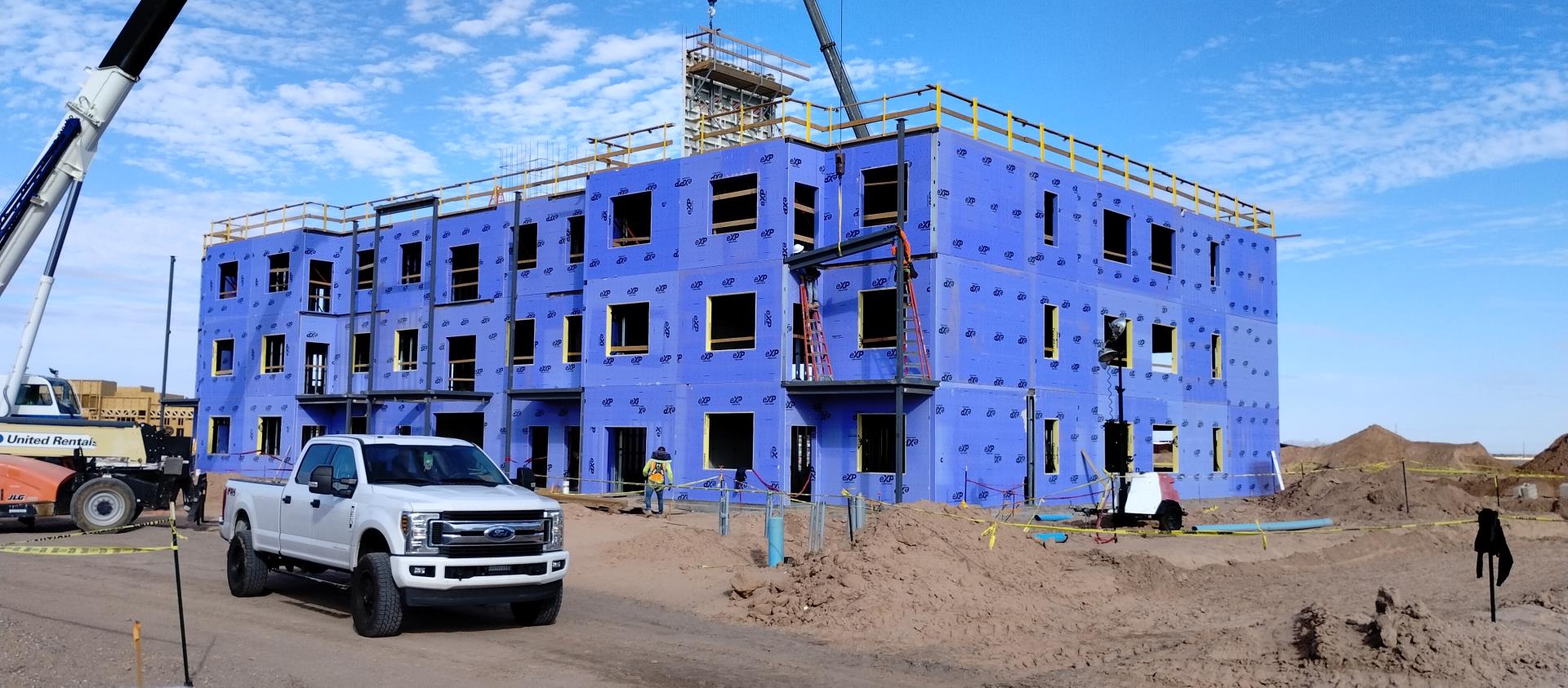 A construction site with a partially built three-story building wrapped in blue sheathing. A white truck and a crane are in the foreground under a clear sky.