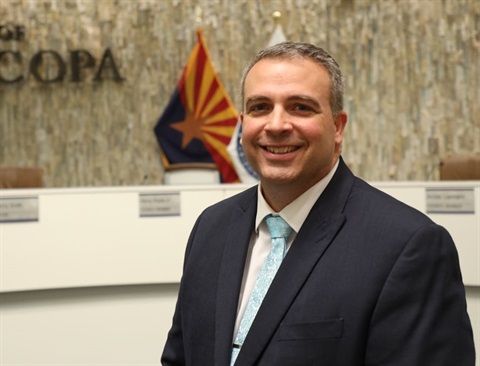 Smiling man in a suit stands in a council chamber, with an Arizona flag and 
