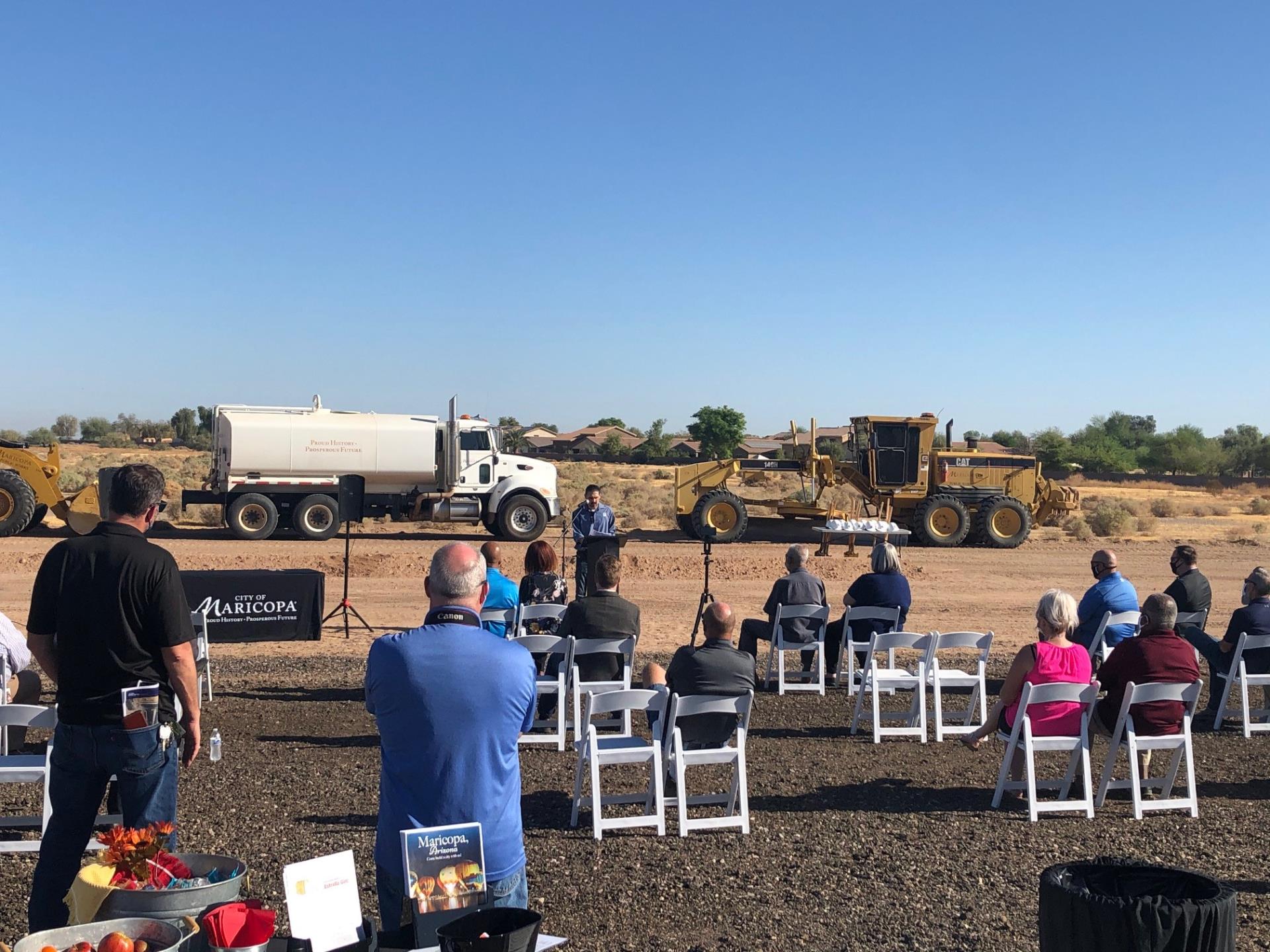 A speaker addresses an audience seated on white folding chairs in a construction site. A water tanker and grader are parked behind them. The atmosphere is formal and businesslike.