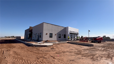 A modern, single-story building under construction on a barren lot under a clear blue sky. Workers and construction equipment are visible on site.