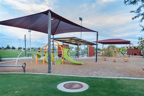 Modern playground with orange and green equipment, shaded by large canopies. Picnic tables under a shelter, all set in a spacious park with a calm ambiance.