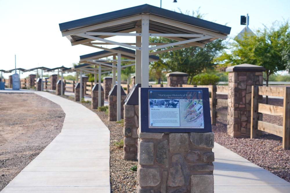 A paved path winds through a historical walk with informational plaques on stone pillars. Metal canopies shade the path, bordered by trees and fencing.