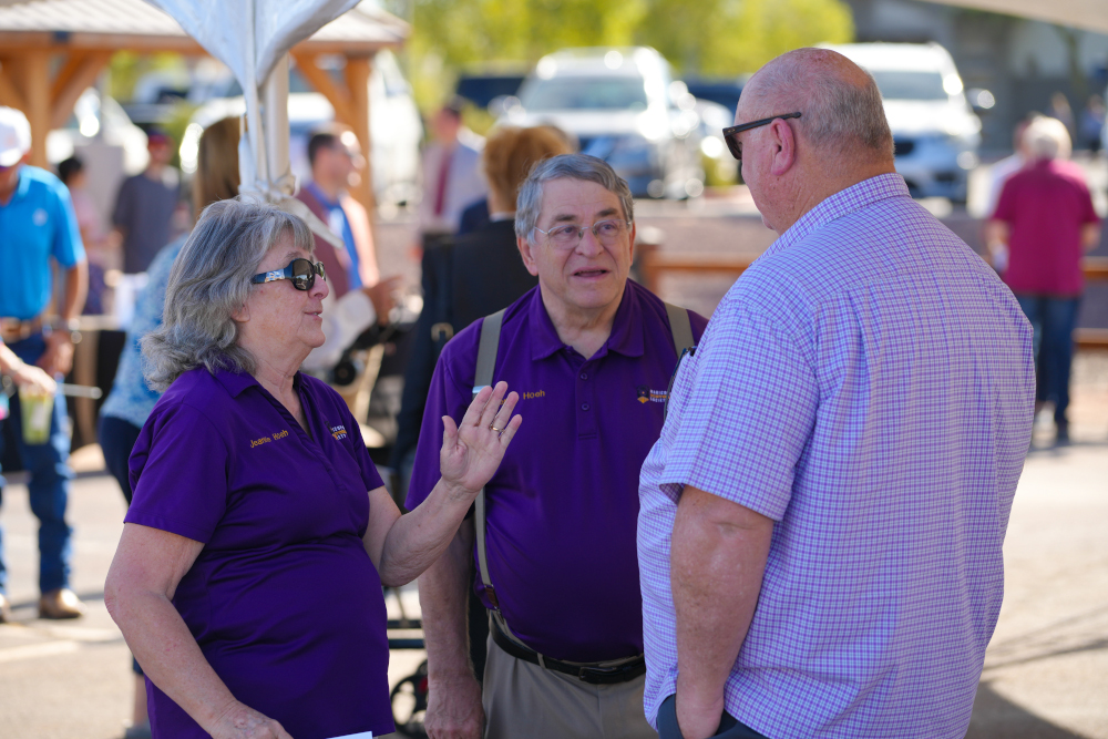 Three people are in conversation outdoors at an event. Two wear purple shirts, engaged and smiling, while the third listens attentively in a checkered shirt.