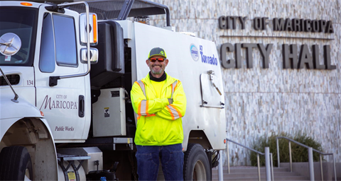 A man in a neon yellow jacket and cap stands smiling with arms crossed in front of a city maintenance truck at Maricopa City Hall.