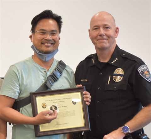 Two people smiling as one holds a framed certificate and badge alongside a police officer in uniform. The atmosphere is celebratory and proud.