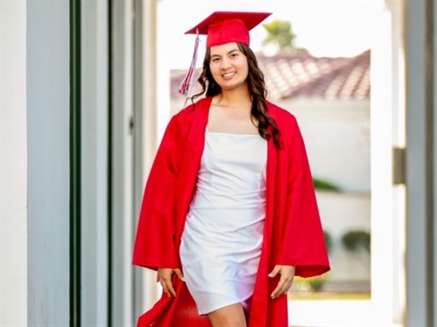 Young woman in red graduation gown and cap, smiling confidently. She wears a white dress underneath. Bright outdoor setting, evokes joy and accomplishment.