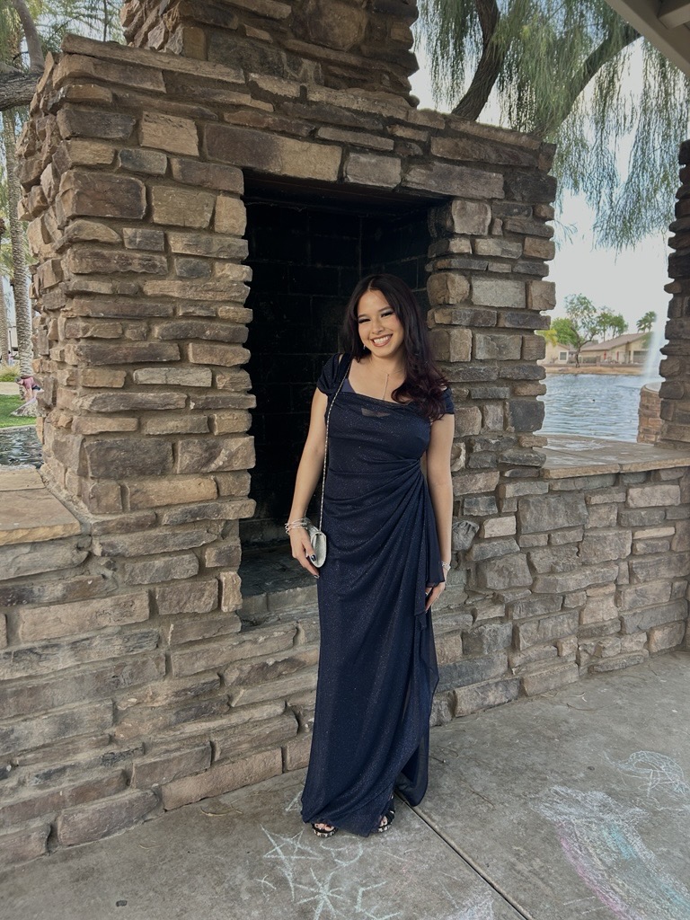 A woman in a navy gown stands smiling beside a stone fireplace outdoors. Chalk drawings appear on the ground, and a lake is visible in the background.